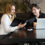 Two young caucasian women meeting, talking, sitting at the table looking in document file or cafe menu, working together beside laptop, copy space